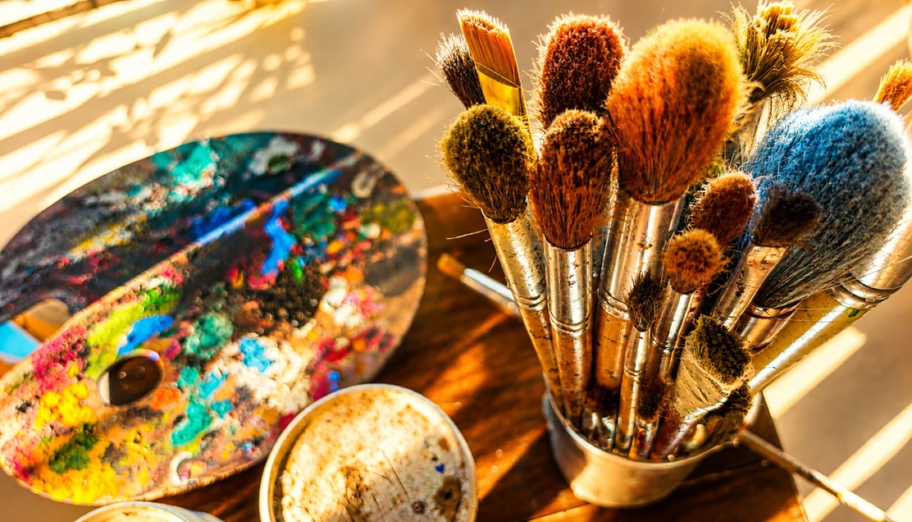 Un bouquet de pinceaux usés dans un pot et une palette couverte de couleurs, posés sur une table en bois dans la lumière rasante du matin.