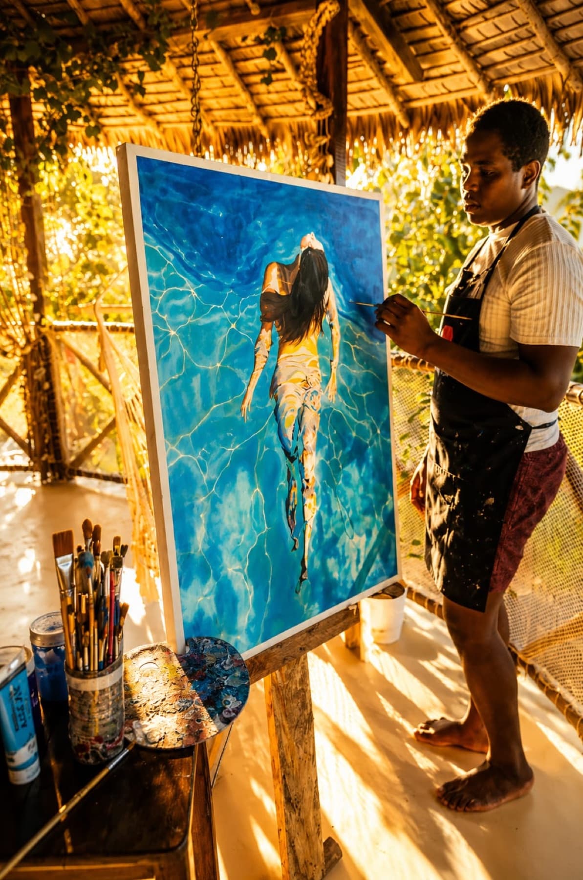 Zago de profil devant son chevalet, occupé à peindre une grande toile bleue, dans son atelier ouvert sur l'extérieur.