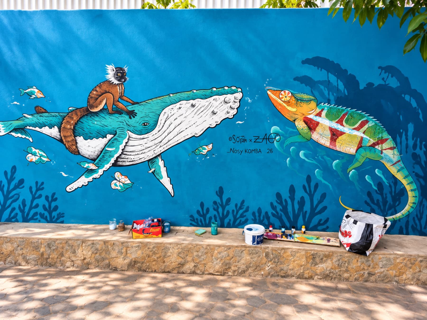 Fresque murale sur fond bleu profond représentant un lémurien chevauchant une baleine à bosse parmi les poissons et les algues, face à un caméléon aux couleurs vives, signée So'ïam et Zago, Nosy Komba.